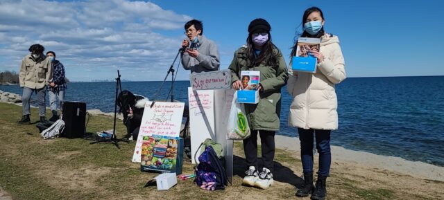 A group of people standing on a beach with signs and a body of water in the background

Description automatically generated with low confidence