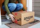 A person bends down to pick up an Amazon box delivered to a doorstep.